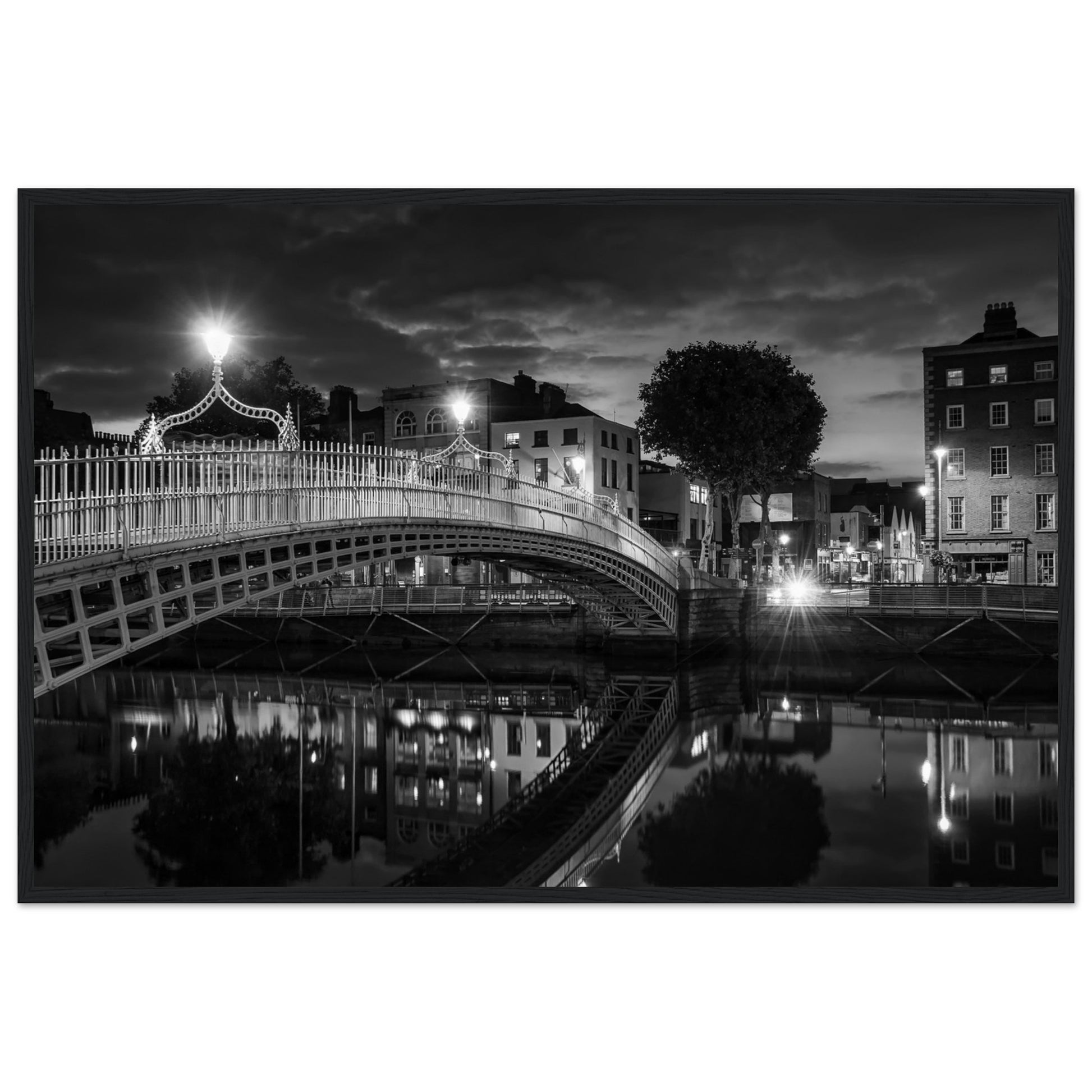 Black and white framed wall art print of the Ha'penny Bridge in Dublin at night. The bridge's historic architecture is beautifully highlighted against the dark sky, with reflections shimmering in the River Liffey below. Ideal for adding elegance to any room or office.