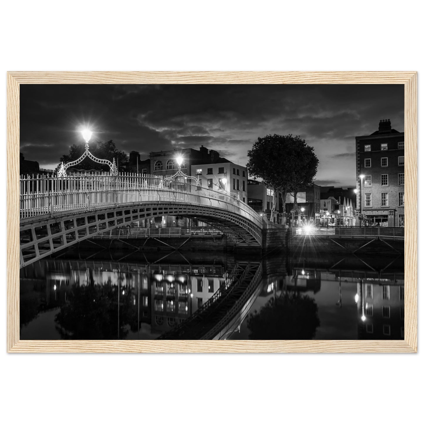 Black and white framed wall art print of the Ha'penny Bridge in Dublin at night. The bridge's historic architecture is beautifully highlighted against the dark sky, with reflections shimmering in the River Liffey below. Ideal for adding elegance to any room or office.