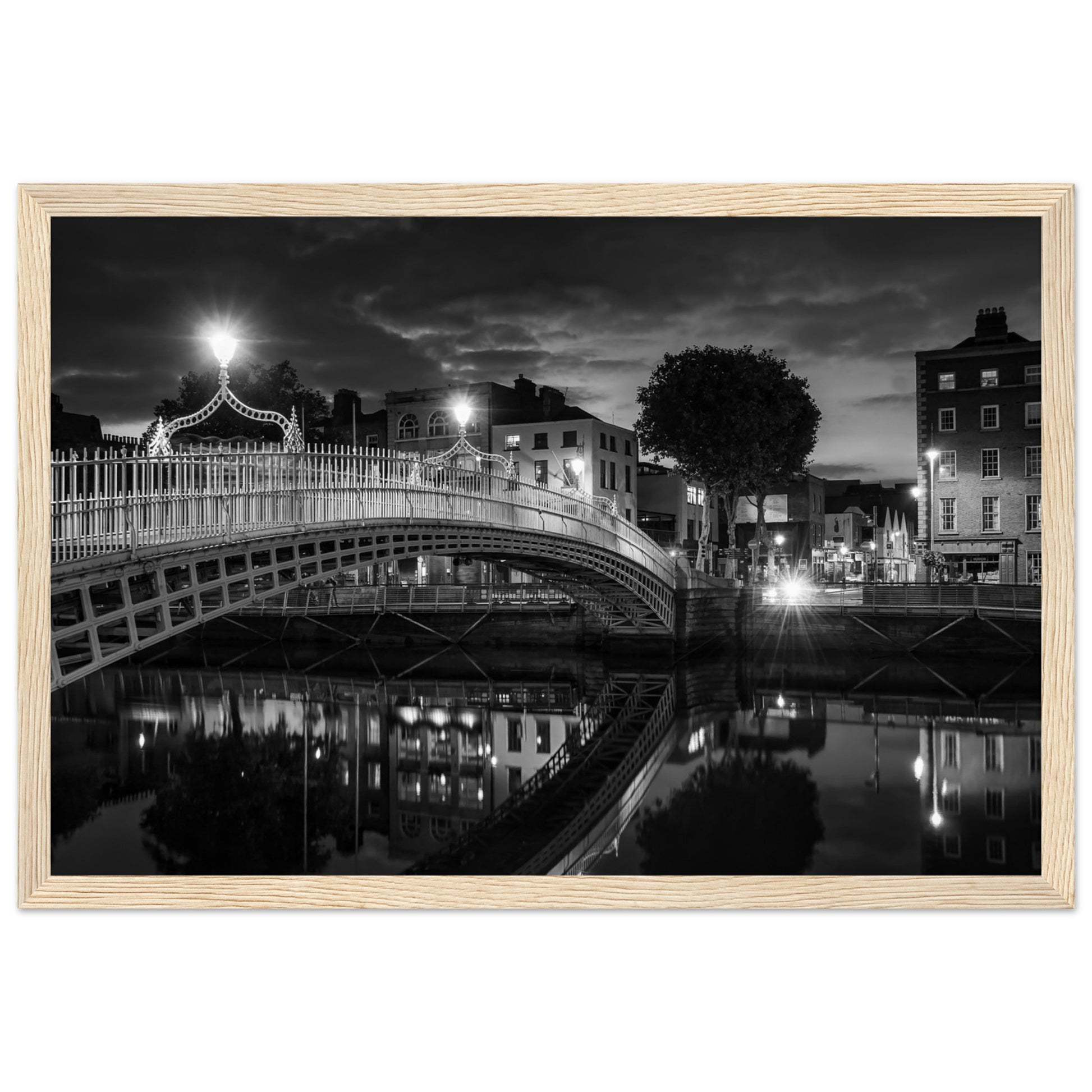 Black and white framed wall art print of the Ha'penny Bridge in Dublin at night. The bridge's historic architecture is beautifully highlighted against the dark sky, with reflections shimmering in the River Liffey below. Ideal for adding elegance to any room or office.