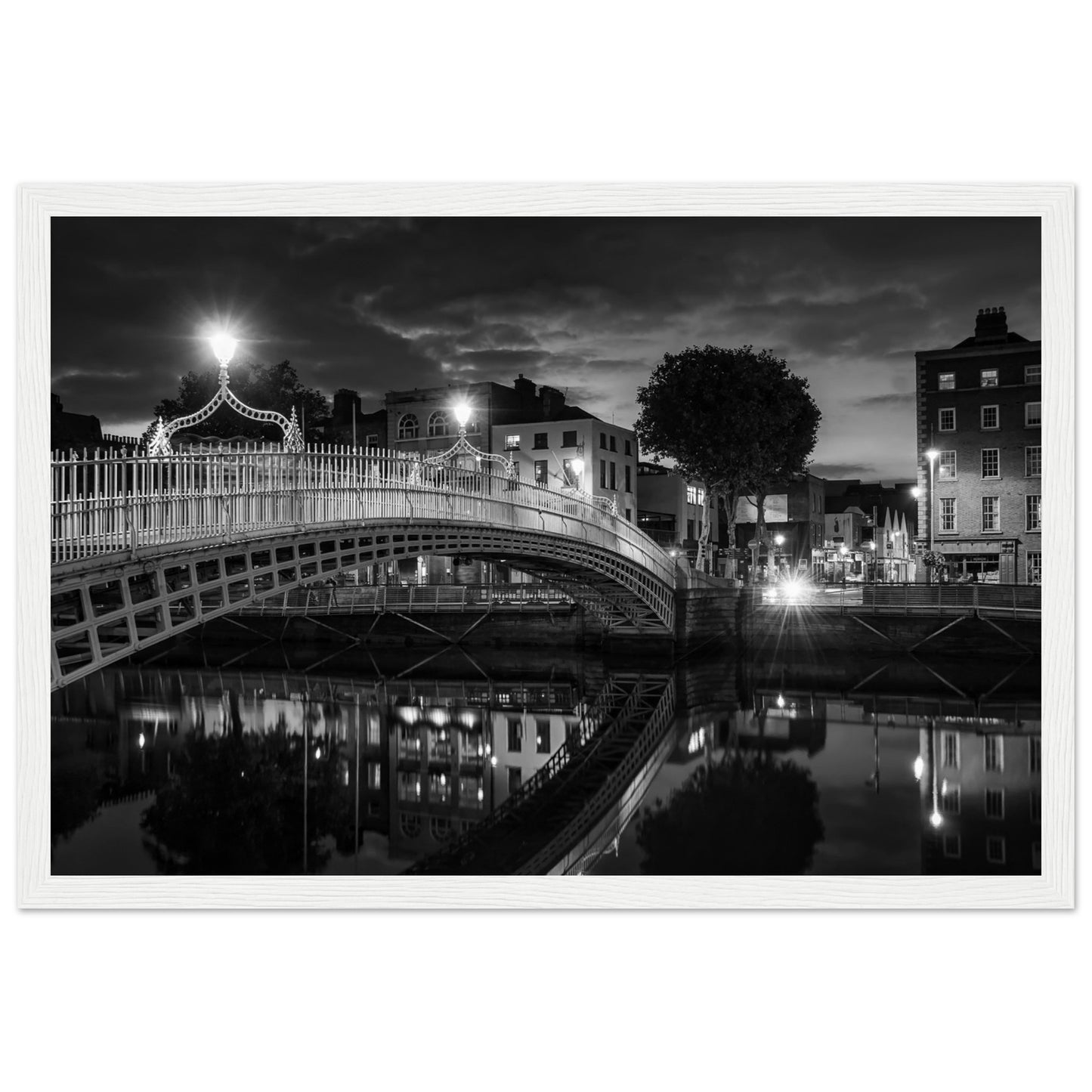 Black and white framed wall art print of the Ha'penny Bridge in Dublin at night. The bridge's historic architecture is beautifully highlighted against the dark sky, with reflections shimmering in the River Liffey below. Ideal for adding elegance to any room or office.