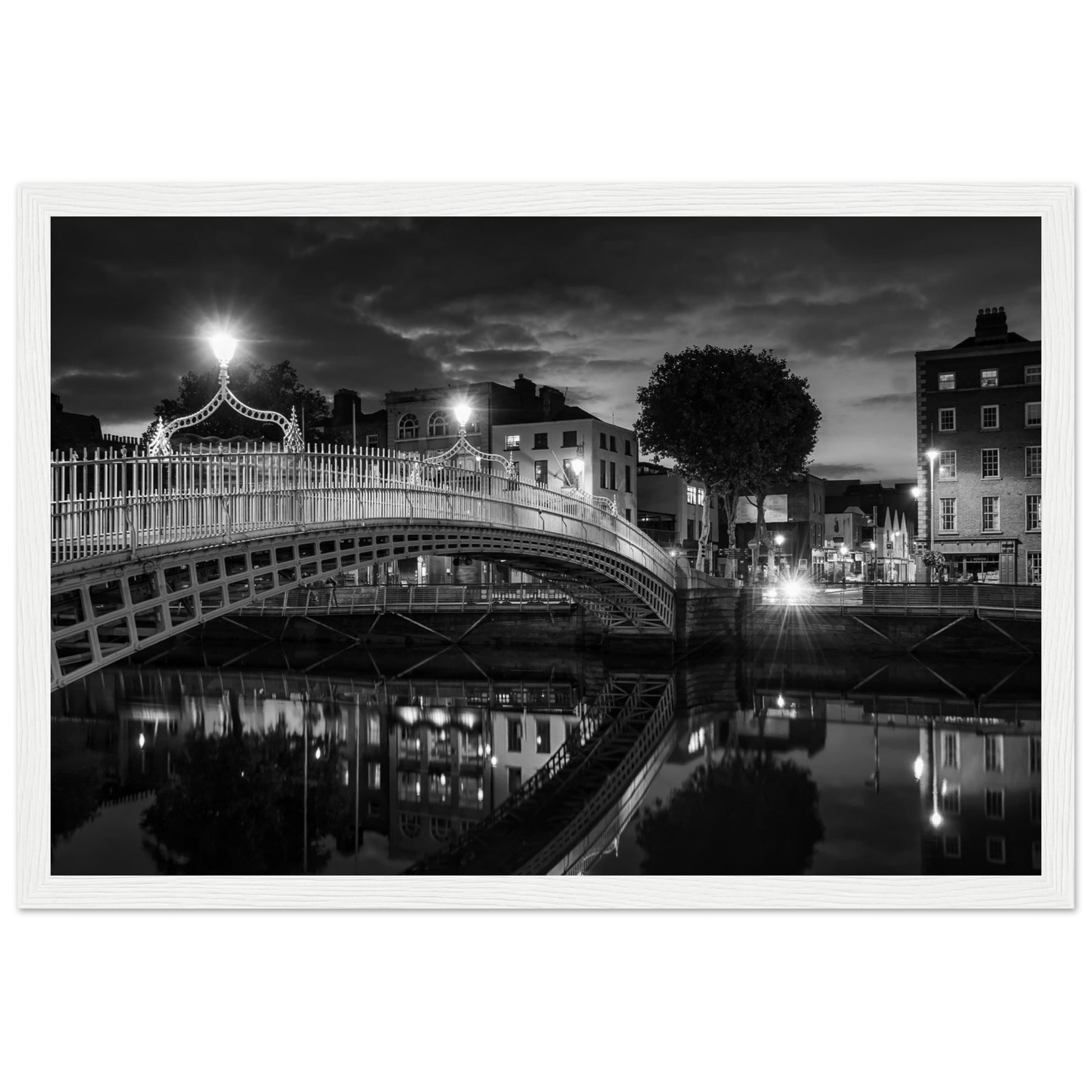 Black and white framed wall art print of the Ha'penny Bridge in Dublin at night. The bridge's historic architecture is beautifully highlighted against the dark sky, with reflections shimmering in the River Liffey below. Ideal for adding elegance to any room or office.