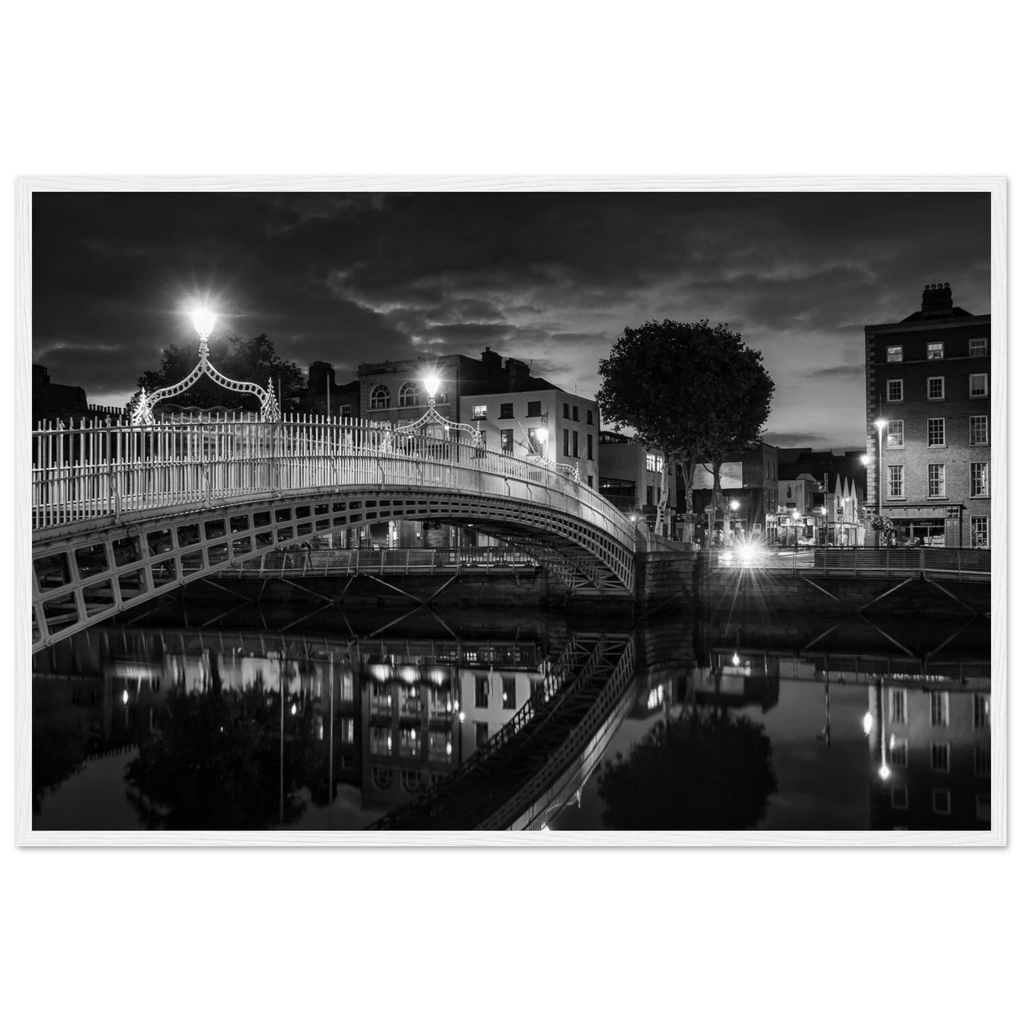 Black and white framed wall art print of the Ha'penny Bridge in Dublin at night. The bridge's historic architecture is beautifully highlighted against the dark sky, with reflections shimmering in the River Liffey below. Ideal for adding elegance to any room or office.