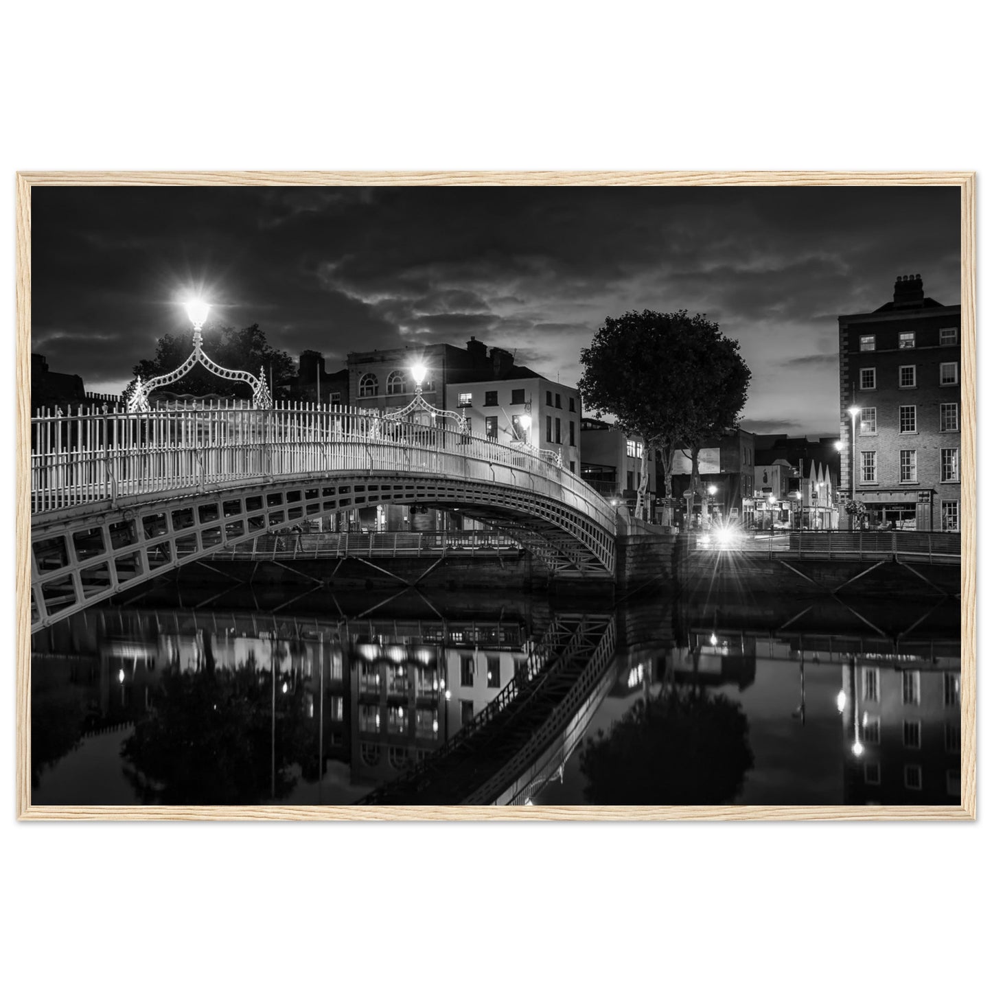 Black and white framed wall art print of the Ha'penny Bridge in Dublin at night. The bridge's historic architecture is beautifully highlighted against the dark sky, with reflections shimmering in the River Liffey below. Ideal for adding elegance to any room or office.