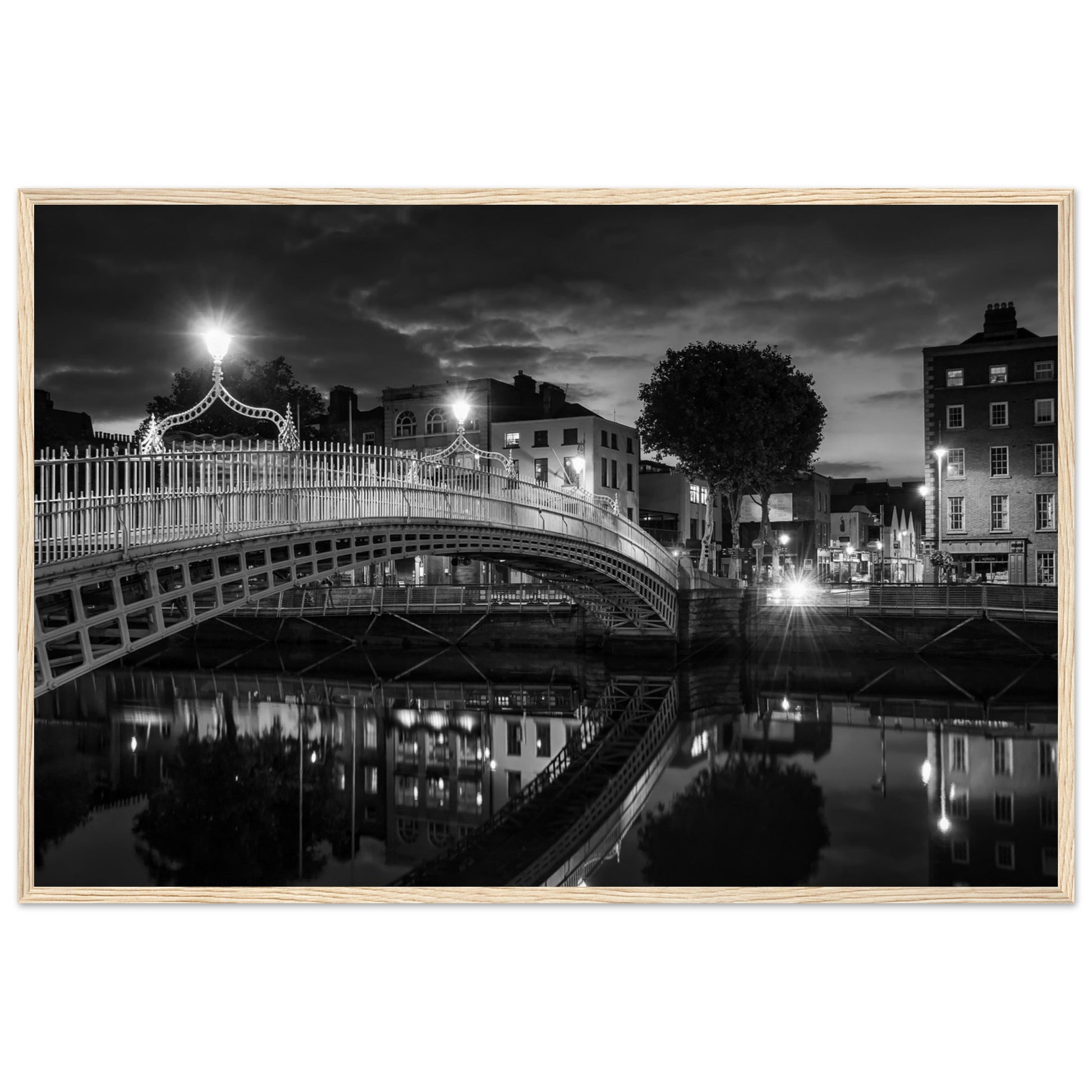 Black and white framed wall art print of the Ha'penny Bridge in Dublin at night. The bridge's historic architecture is beautifully highlighted against the dark sky, with reflections shimmering in the River Liffey below. Ideal for adding elegance to any room or office.