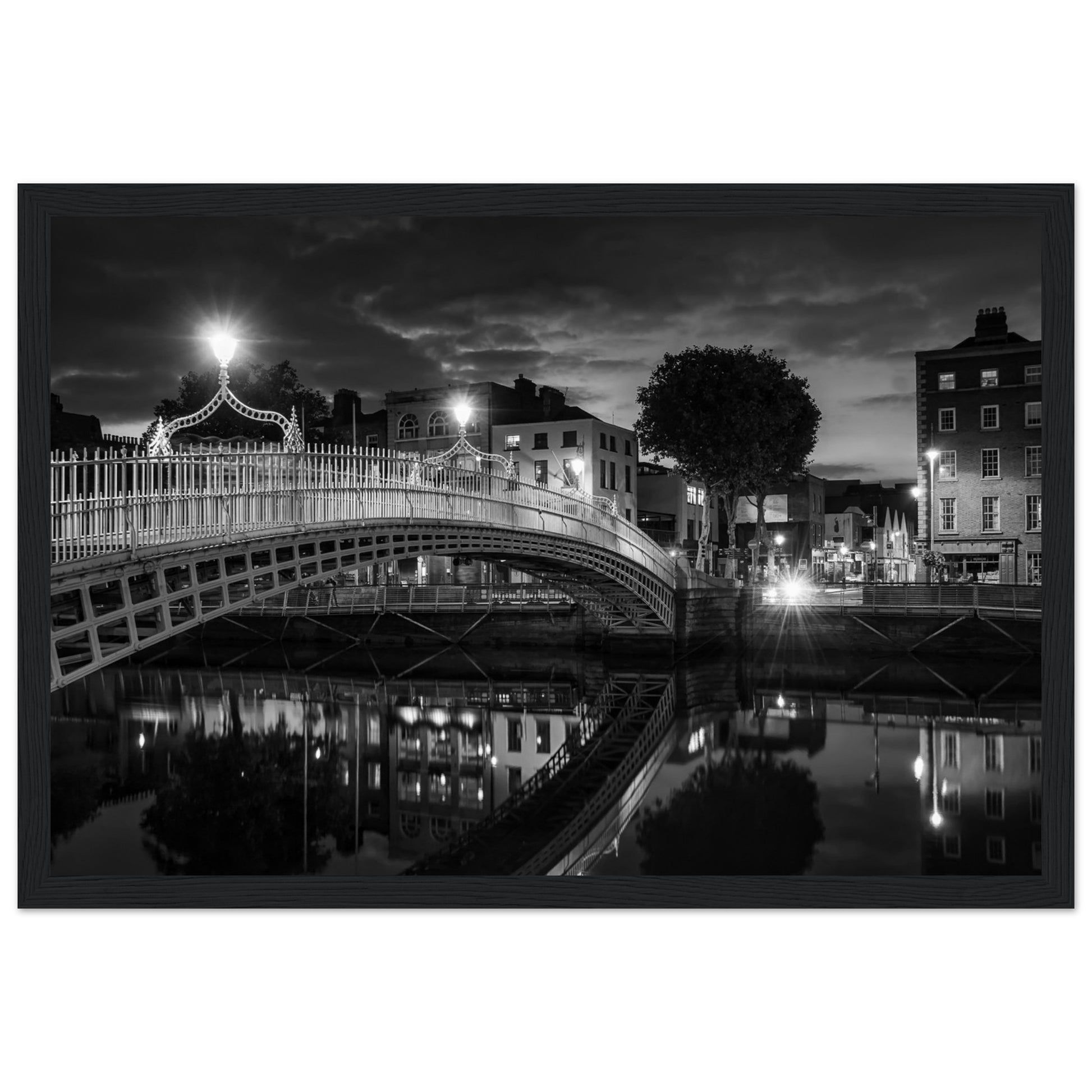 Black and white framed wall art print of the Ha'penny Bridge in Dublin at night. The bridge's historic architecture is beautifully highlighted against the dark sky, with reflections shimmering in the River Liffey below. Ideal for adding elegance to any room or office.