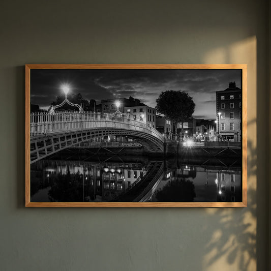 Night View of Ha'penny Bridge framed print in a natural oak frame on a dark green wall beside a window, dappled plant shadow