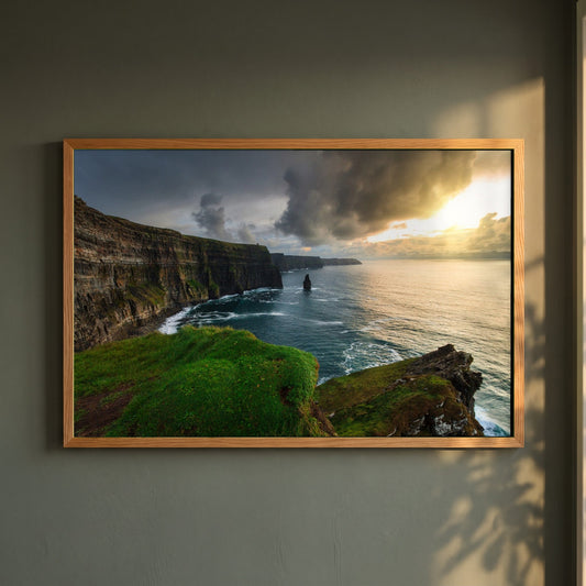 Cliffs of Moher framed print in a natural oak frame on a dark green wall beside a window, dappled plant shadow