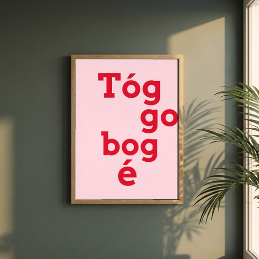 Tóg é go bog é framed print in a natural oak frame on a dark green wall beside a window, dappled plant shadow