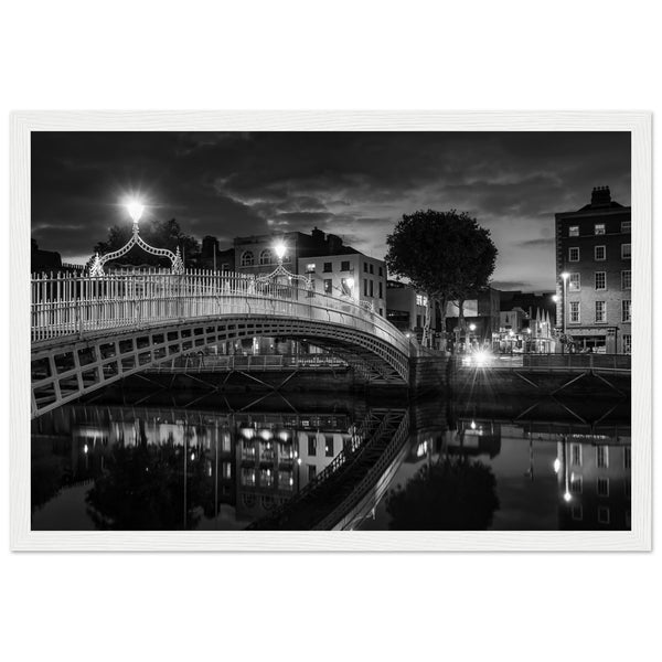  Black and white framed wall art print of the Ha'penny Bridge in Dublin at night. The bridge's historic architecture is beautifully highlighted against the dark sky, with reflections shimmering in the River Liffey below. Ideal for adding elegance to any room or office.