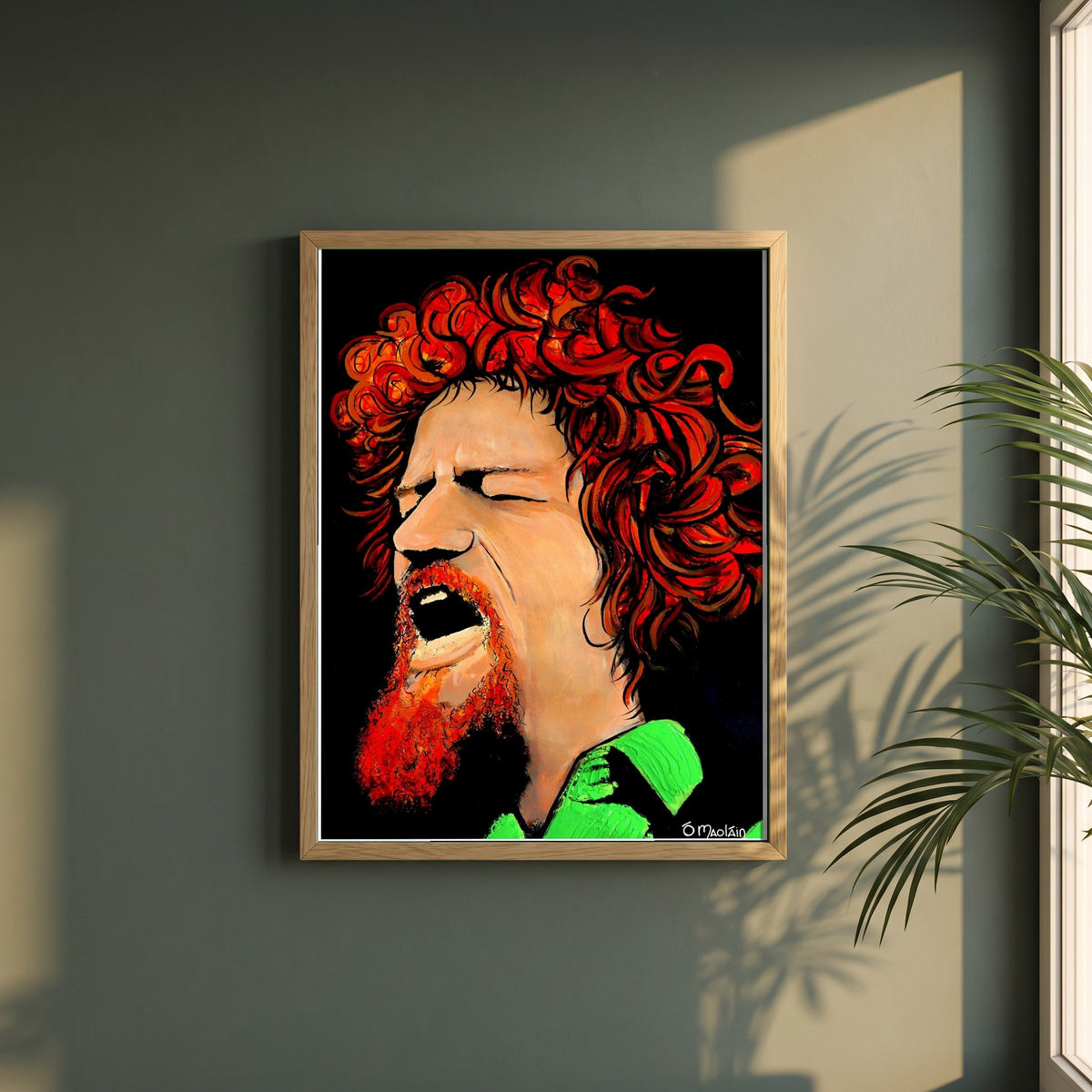 Luke Kelly framed print in a natural oak frame on a dark green wall beside a window, dappled plant shadow