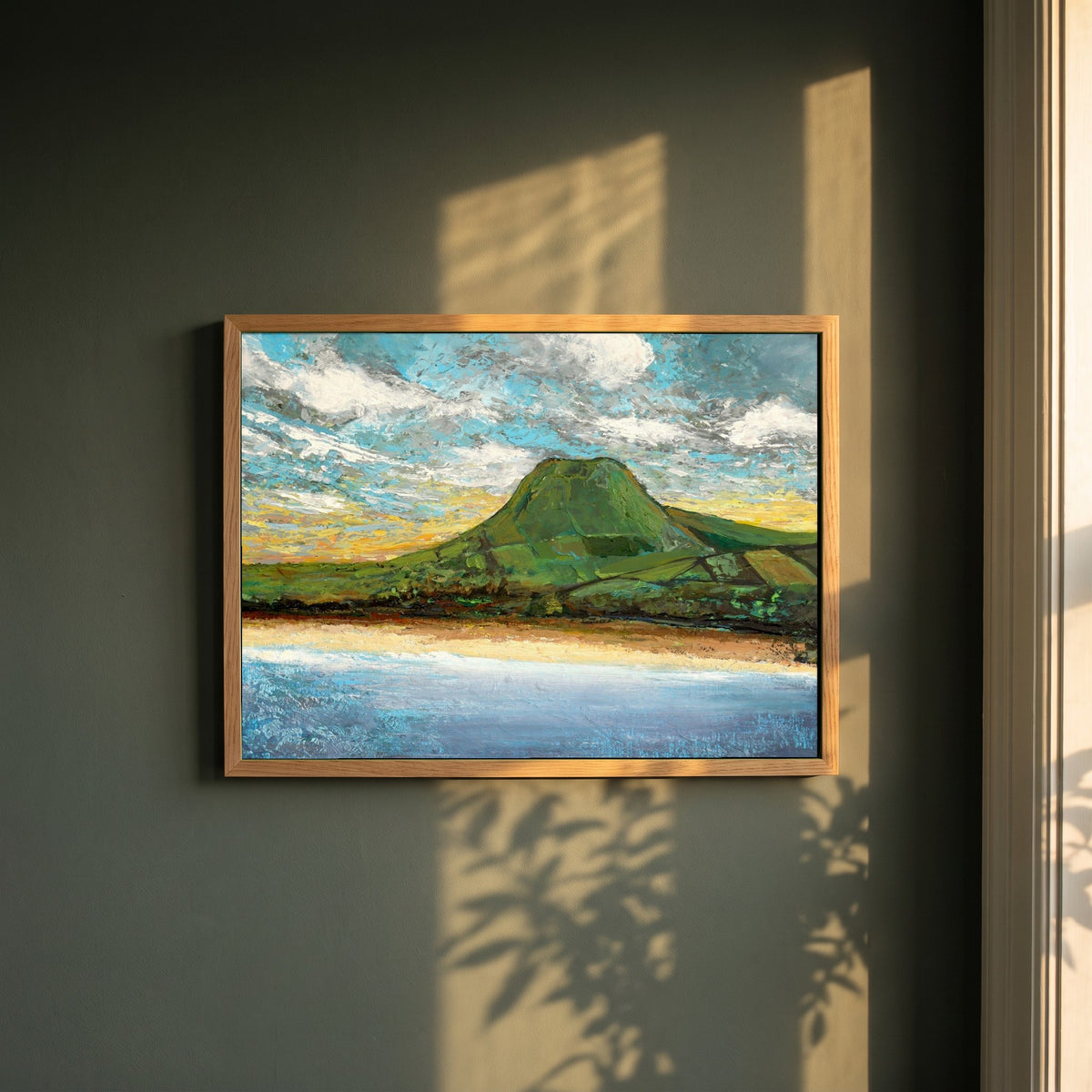 Lurig Mountain at Sunset framed print in a natural oak frame on a dark green wall beside a window, dappled plant shadow