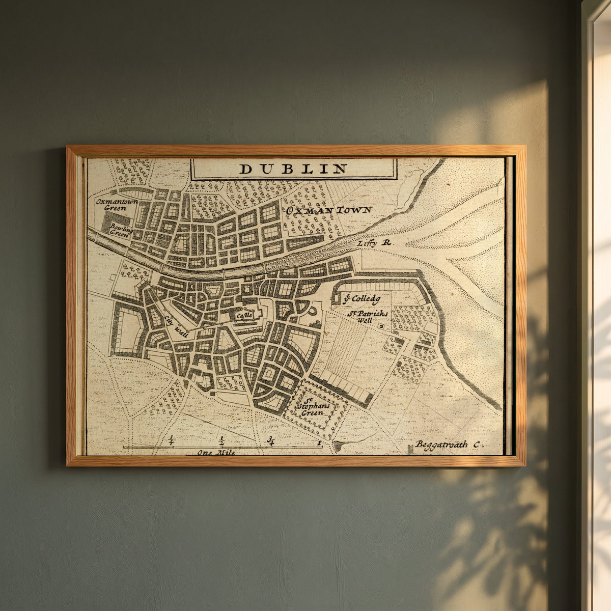 Map of Dublin City in 1714 framed print in a natural oak frame on a dark green wall beside a window, dappled plant shadow