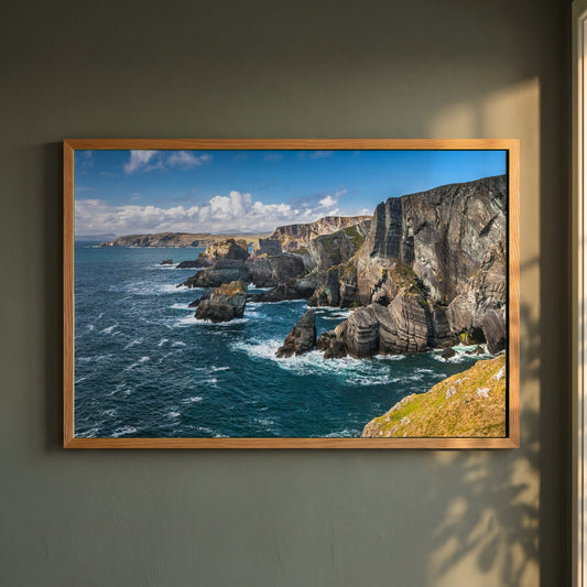 Mizen Head Ireland framed print in a natural oak frame on a dark green wall beside a window, dappled plant shadow