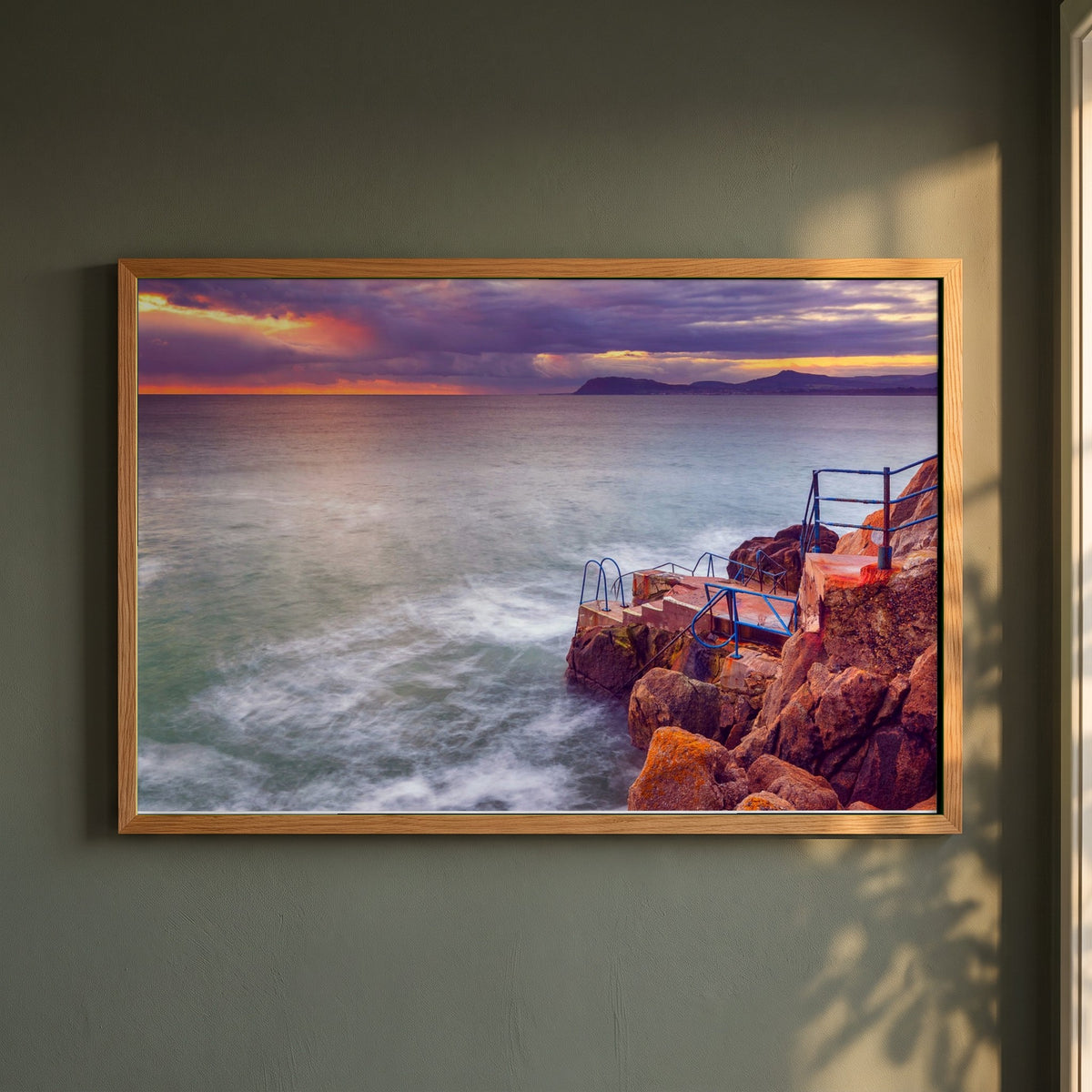 Vico Swimming Dalkey framed print in a natural oak frame on a dark green wall beside a window, dappled plant shadow
